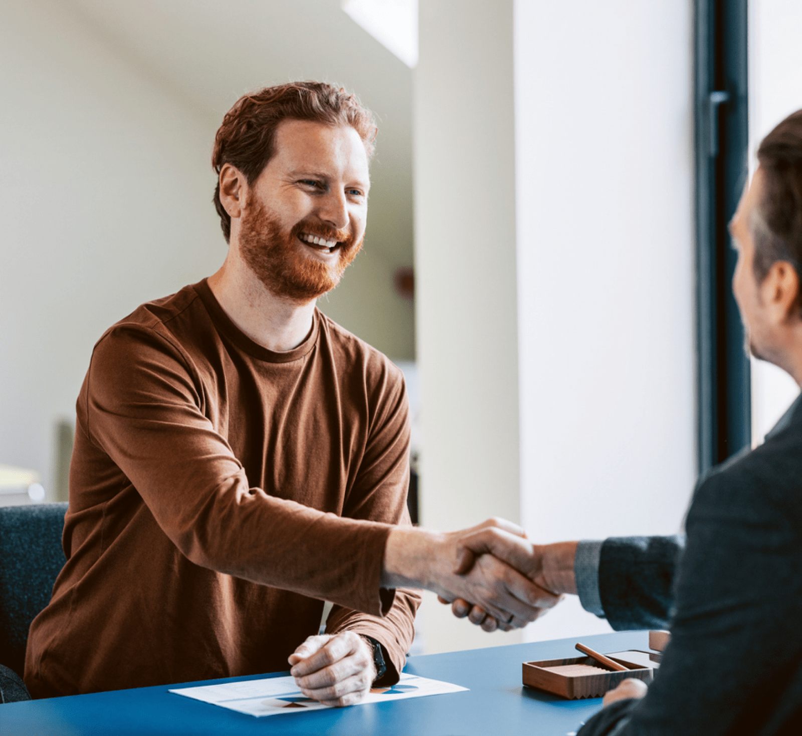 Smiling bearded man in a brown shirt shakes hands with a suited professional across a desk, conveying a warm, successful business meeting.