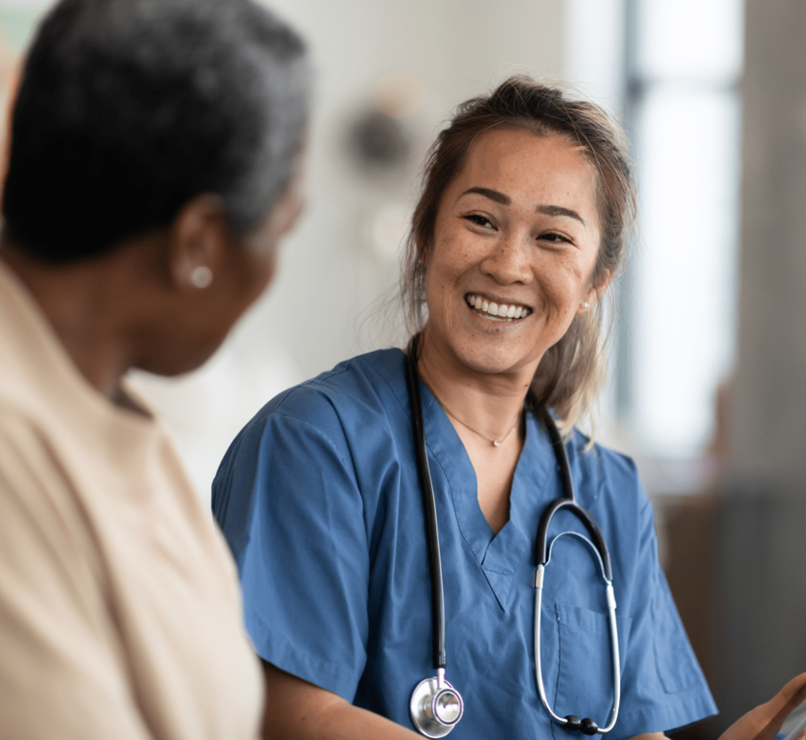 Smiling Asian healthcare professional in blue scrubs and stethoscope speaks warmly with an older Black patient in a bright clinic.