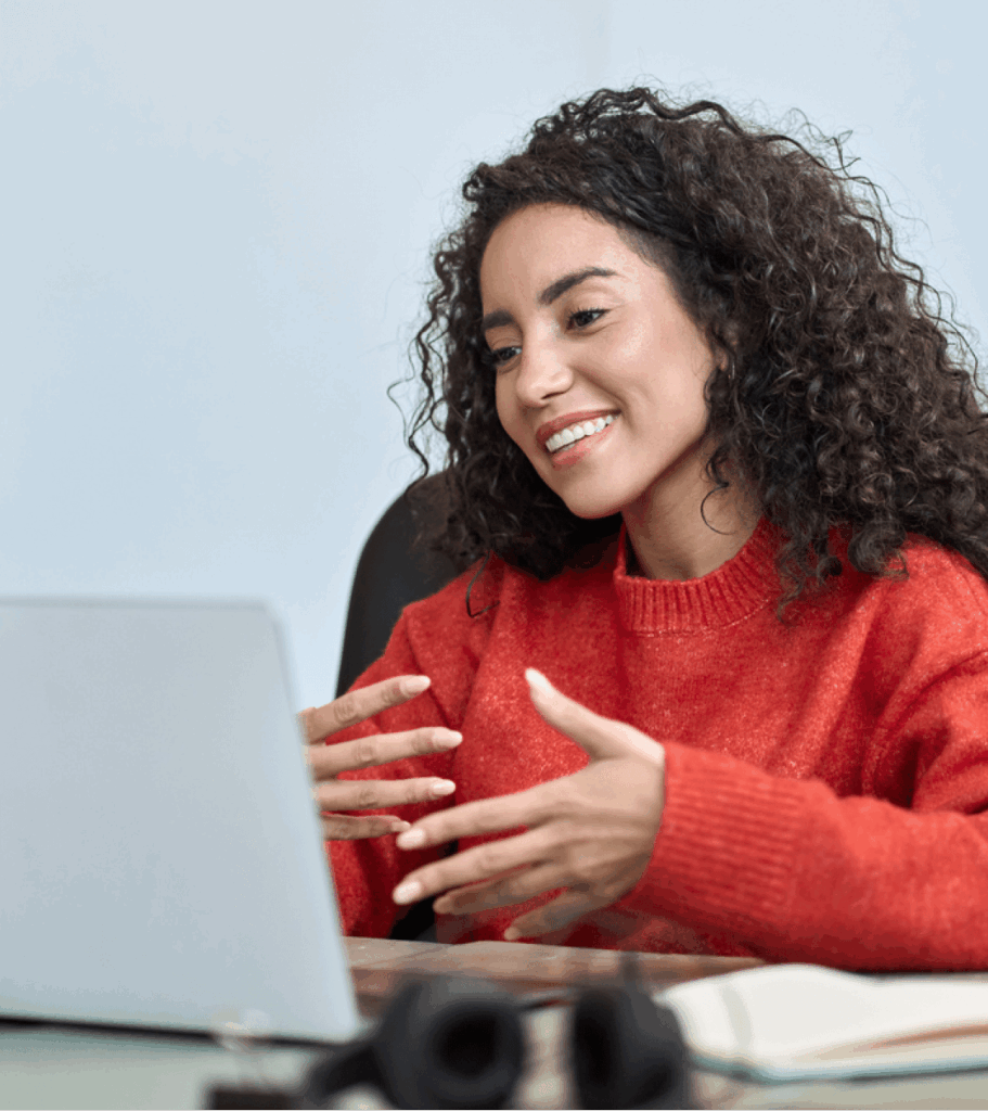 Smiling woman with curly hair in a red sweater gestures beside a laptop during a friendly video call, creating a warm, engaged mood.