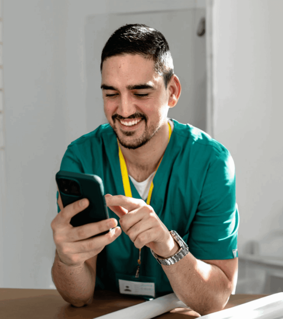 Smiling healthcare worker in teal scrubs and ID badge uses a smartphone at a desk in bright light, conveying warmth and engagement.