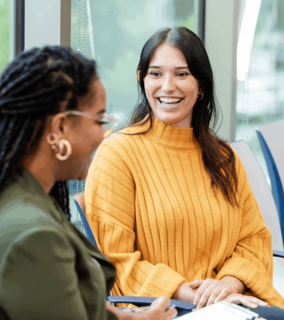 Two women chat in a bright office: a smiling woman in a mustard sweater faces a colleague with locs and glasses, conveying warmth and teamwork.